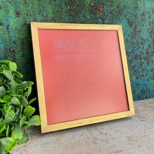 Peut inclure: Un cadre photo carré avec une fine bordure métallique dorée et un insert rouge uni. L'insert présente un texte blanc pâle indiquant EPA, RAM et 12,5x12,5. Le cadre a un design simple et épuré avec des coins nets et un profil plat.