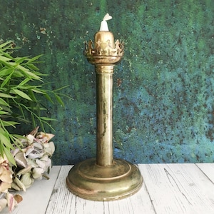 May include: A vintage brass oil lamp with a tall, slender stem and a round base. The top has a decorative crown design and a white wick. The lamp is set against a textured green and blue background.