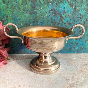 May include: A silver-coloured, footed bowl with two handles. The interior has a gold-coloured finish. The bowl has a decorative leaf pattern etched around the rim. The bowl is likely used for serving.