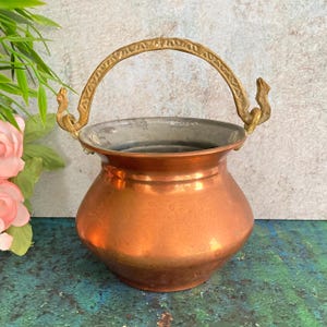 Puede incluir: Una pequeña olla de cobre antigua con un asa de latón. La olla tiene un cuerpo redondeado y un borde ancho. El asa es arqueada y está unida a la olla con soportes decorativos. El cobre tiene un color marrón rojizo cálido.