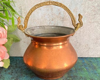 Puede incluir: Una pequeña olla de cobre antigua con un asa de latón. La olla tiene un cuerpo redondeado y un borde ancho. El asa es arqueada y está unida a la olla con soportes decorativos. El cobre tiene un color marrón rojizo cálido.