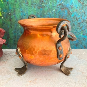 以下が含まれることがあります： 丸みを帯びたボディと3つの装飾的な足を持つ、小さなハンマー加工された銅製の鍋。鍋には2つの濃い色の金属製のハンドルがあり、装飾目的で使用される可能性があります。銅は古びたオレンジブラウンの緑青を持っています。