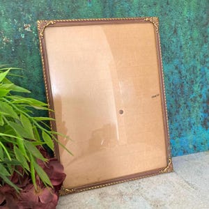 May include: A rectangular picture frame with a thin, dark brown metallic border featuring decorative gold-toned corner accents. The frame contains a clear glass pane covering a plain, light tan background surface with a subtle grid pattern.