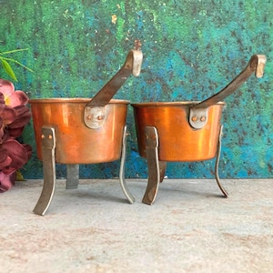 Op de afbeelding: Twee koperen potten met metalen handvatten en elk drie poten. De potten hebben een gepolijste oranjebruine afwerking en zijn bevestigd met zichtbare klinknagels. Het ontwerp bevat een lange steel aan de zijkant van elke kom.