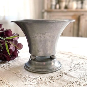 May include: A silver-toned metal trophy cup with a wide, flared rim and a circular base supported by three small spherical feet. The front of the cup features the engraved text "IK SPORT HEDERSPRIS" centered on the smooth, metallic surface.