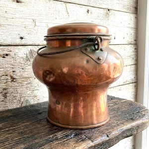 Op de afbeelding: Een ronde koperen container met een deksel en een donkere, verweerde metalen zwenkhendel bevestigd aan driehoekige platen. Het koperen oppervlak heeft een gehamerde textuur met zichtbare deuken en een warme, roodbruine afwerking.