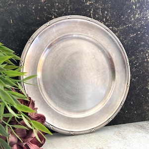 May include: A round silver-toned metal plate with a smooth, reflective centre surface and a decorative rim featuring a repeating etched cross-hatch pattern along the outer edge. The plate has a polished finish with subtle signs of wear.