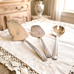 Puede incluir: Un juego de tres utensilios de servicio de metal plateado que incluye una pala para pasteles, una espátula pequeña y una cuchara ranurada. Cada mango está decorado con un patrón floral en relieve repetido a lo largo de los bordes.