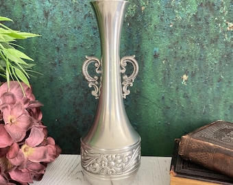 May include: A silver vase with decorative handles and a floral pattern around the base. The vase has a narrow neck and a flared base. The background is a mottled green and brown colour.