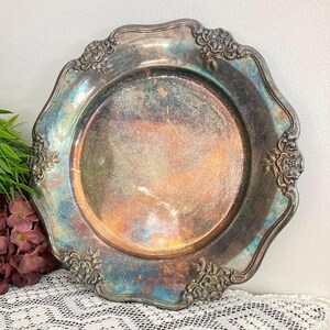 May include: A round silver-plated serving tray with a scalloped edge and embossed floral details. The surface displays a natural patina with iridescent blue, purple, and copper-toned oxidation marks across the center and the decorative rim.