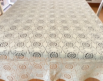 Könnte beinhalten: Cremefarbene, gehäkelte Tischdecke mit sich wiederholendem Blumenmuster. Die rechteckige Tischdecke bedeckt einen Holztisch. Das komplizierte Design weist durchbrochene Details auf, die ein zartes und strukturiertes Aussehen erzeugen. Die Gesamtästhetik ist Vintage und handgefertigt.