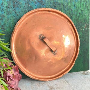Könnte beinhalten: Ein runder Kupfertopfdeckel mit Griff. Der Deckel hat eine leicht gehämmerten Textur und eine warme, rotbraune Farbe. Der Griff ist ein einfacher Riemen, der mit Nieten befestigt ist.