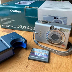 May include: A silver Canon Digital IXUS 400 camera with a wrist strap, a 1GB Silicon Power CompactFlash memory card, a dark blue battery charger, and the original product box featuring the text DIGITAL IXUS 400 and Canon branding.