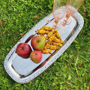 Puede incluir: Una bandeja de servir plateada con un borde de cuentas, decorada con tres manzanas rojas y un montón de caramelos envueltos en oro. Dos vasos con tallo vacíos están en la bandeja.