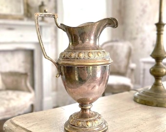 May include: A metallic jug with a tarnished silver and copper finish, featuring a curved handle, a wide spout, and a pedestal base. The body is decorated with a band of embossed circular motifs around the neck and the base.