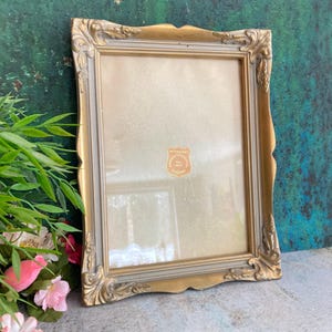 May include: A rectangular picture frame with a gold-toned finish and ornate, carved corner details. The frame features a light gray inner border and a glass front containing a small, shield-shaped sticker with the text AKTIEBOLAGET TEL 1881.