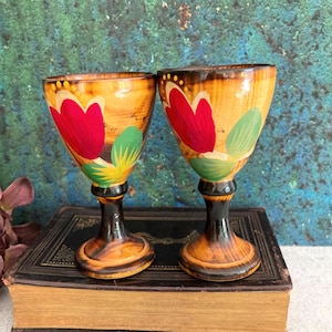Könnte beinhalten: Zwei Holzbecher mit Tulpendesign. Die Becher haben einen hellbraunen Sockel mit einem dunkelbraunen Stiel und Rand. Jeder Becher zeigt eine rote Tulpe mit grünen Blättern und gelben Akzenten. Die Becher werden auf einem Vintage-Buch ausgestellt.