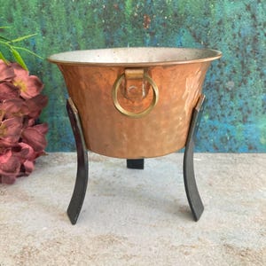 Può includere: Una ciotola in rame martellato con un manico ad anello in ottone, sostenuta da un supporto in metallo nero. La ciotola ha un bordo largo e una superficie strutturata. Il supporto ha tre gambe curve. Design vintage e decorativo.