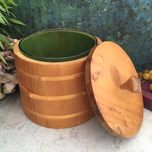 Op de afbeelding: Een houten ijsemmer met een groene binnenkant en een bijpassend houten deksel. De emmer is gemaakt van lichtgekleurd hout met horizontale banden. Het deksel heeft een kleine, vierkante handgreep. De emmer is open en onthult de groene binnenkant.