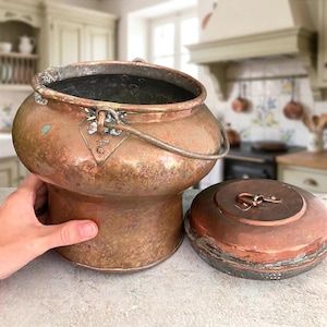 May include: A round copper pot with a wide opening, a curved body, and a metal swing handle attached by triangular brackets. The pot features a weathered patina and is accompanied by a separate, matching circular copper lid with a small handle.