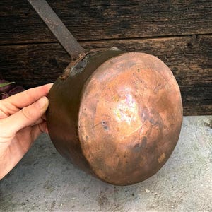 Puede incluir: Un cazo de cobre redondo con una superficie desgastada y oxidada, que cuenta con un mango largo y plano de metal oscuro sujeto con un remache visible. El exterior muestra signos de antigüedad, incluyendo manchas oscuras y una pátina mate.