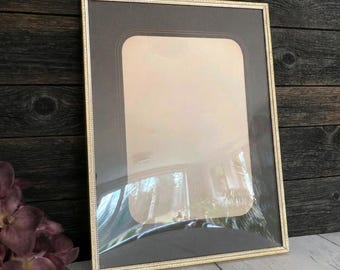 May include: A rectangular picture frame with a thin gold-toned metallic border and a dark brown matting. The frame features a rounded rectangular inner opening, creating a layered appearance. The glass surface reflects light and indoor surroundings.
