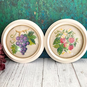 Puede incluir: Dos obras de arte redondas en punto de cruz enmarcadas de color crema. Una presenta un racimo de uvas moradas con hojas verdes, la otra un ramo de fresas rojas y flores blancas. Ambas están sobre un fondo de tela beige.