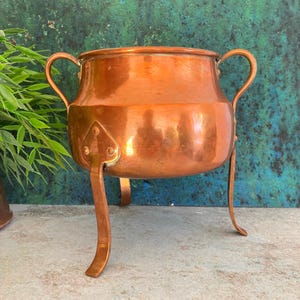 May include: A polished copper pot with a rounded body, two curved side handles, and three slender legs attached with triangular plates. The metallic surface reflects light, showing a warm orange-brown hue and a smooth, rounded rim.