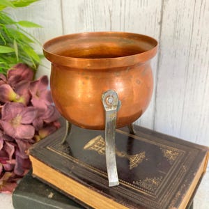 May include: A small, antique copper cauldron with a rounded body and a sturdy metal handle. The cauldron sits on three small legs and is displayed on top of a stack of vintage books. The copper has a warm, reddish-brown hue.