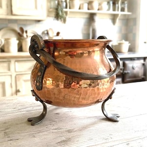Peut inclure: Un chaudron en cuivre martelé de forme ronde avec une large ouverture et une finition métallique polie. Le récipient possède une anse en fer sombre et robuste fixée par des rivets et repose sur trois pieds en fer courbés fixés à la base.