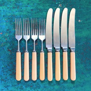 May include: A set of vintage silverware, including forks and knives, arranged on a teal surface. The forks have four tines and the knives have a rounded blade. The handles are a light cream color, offering a classic aesthetic.