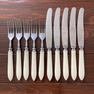 May include: A set of vintage silverware, including forks and knives, arranged on a dark wooden surface. The forks and knives have silver blades and off-white handles. The arrangement suggests a formal dining setting.