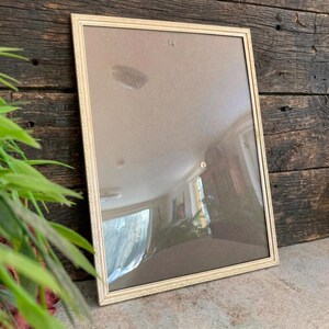 May include: A rectangular picture frame with a thin, metallic gold-toned border featuring a delicate, repeating embossed pattern along the edges. The frame contains a clear glass pane, reflecting the surrounding indoor environment.