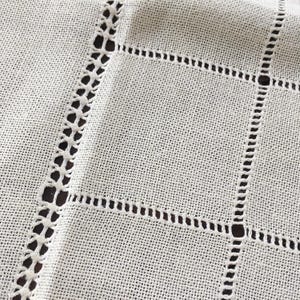 May include: Close-up of a white linen fabric with a grid pattern. The grid is formed by a combination of openwork and stitched lines, creating a textured and decorative surface. The fabric appears to be a tablecloth or similar textile.