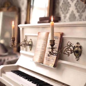 Pode incluir: Um piano vertical branco apresenta dois candeeiros de parede em latão ornamentado montados no painel frontal, cada um com uma vela branca acesa. Um livro de música antigo e aberto com notação musical repousa na estante do piano.