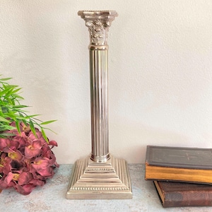 May include: Silver-coloured, fluted column candle holder with a square base and ornate capital. The decorative pillar is approximately 30 cm tall. The design is reminiscent of classical architecture, suitable for home decor.