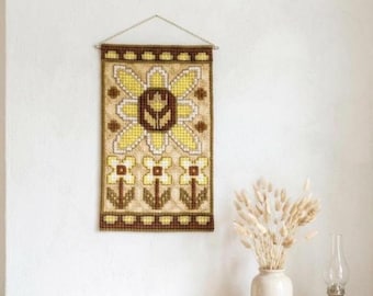 Puede incluir: Un tapiz rectangular con un patrón de punto de cruz que muestra una flor central grande sobre tres flores más pequeñas. El diseño utiliza tonos amarillos, marrones y crema sobre una tela texturizada, suspendido por una fina cadena metálica.