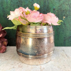 May include: A metallic, barrel-shaped container with a polished silver-tone finish and horizontal ribbed detailing, filled with a bouquet of artificial light pink flowers featuring green leaves and a single small, closed bud at the top center.
