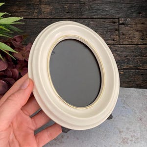 May include: An oval-shaped picture frame with a smooth, off-white finish and a thin, metallic gold-colored inner rim. The frame features a stepped, tiered border design and a dark, solid-colored center insert, presented in a vertical orientation.