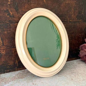 May include: An oval picture frame with a cream-colored border and a thin gold inner rim. The frame contains a green insert with the text 10x14 printed in gold. The frame features a smooth, rounded edge design and a clean, simple construction.