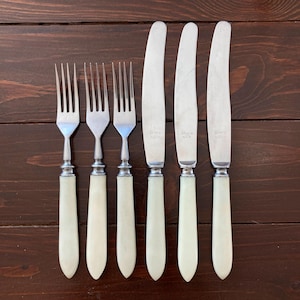 Peut inclure: Un ensemble de couverts anciens, comprenant trois fourchettes et trois couteaux, disposés sur une surface en bois foncé. Les couverts présentent des lames et des dents argentées avec des manches blanc cassé. Les couteaux ont une lame incurvée.