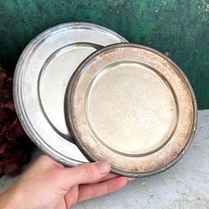 May include: Two round silver-toned metal plates with a decorative cross-hatch pattern along the rim. The larger plate has a polished finish, while the smaller plate shows significant tarnishing and discoloration around the outer edge.