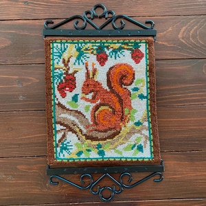 May include: A framed needlepoint featuring a squirrel. The squirrel is depicted in shades of orange and brown, perched on a branch with red berries and green foliage. The frame is dark brown with a decorative black metal hanger.