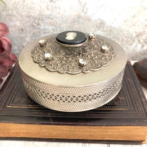 May include: A round silver-toned metal trinket box featuring an intricate filigree lid adorned with four clear rhinestones and a central black circular inlay with a white cameo profile. The sides of the box have a textured, woven metal pattern.