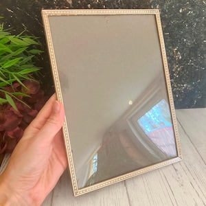 Puede incluir: Un marco de fotos rectangular plateado con un borde decorativo. El marco tiene un respaldo gris y un frente de vidrio. El marco está sostenido por una mano, lo que sugiere su tamaño y uso para exhibir fotografías u obras de arte.