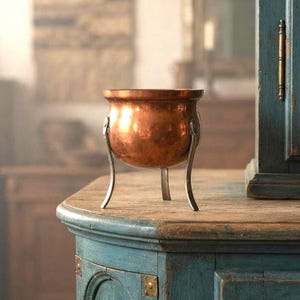 May include: A small, round, polished copper pot with a wide opening and a smooth, reflective surface. The pot is supported by three slender, curved metal legs that are attached to the sides of the vessel, elevating it above the surface.