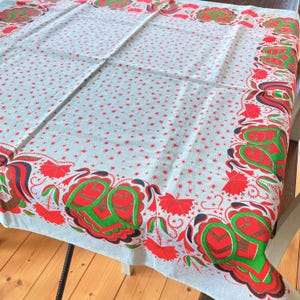 May include: A white tablecloth with a red floral border and red star-shaped accents. The border features stylized red and green floral designs with black accents. The tablecloth is spread over a wooden table, showcasing its decorative pattern.