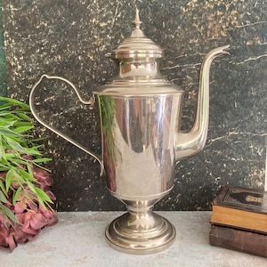 Könnte beinhalten: Eine silberfarbene antike Kaffeekanne mit einem langen, gebogenen Ausguss und einem Griff. Die Kanne hat einen Deckel mit dekorativer Spitze und steht auf einem Sockel. Der Hintergrund ist eine dunkle, marmorierte Oberfläche.