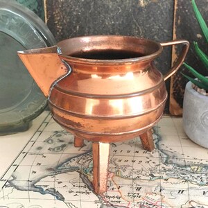 May include: A copper pitcher with a rounded body, a wide spout, and a single handle. The vessel features three short, straight legs that elevate the base. The metallic surface shows a warm, polished finish with subtle signs of age and wear.