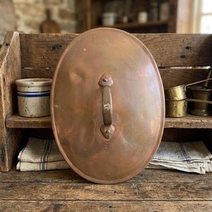 Peut inclure: Un couvercle ovale en cuivre doté d'une poignée centrale en métal portant l'inscription gravée JPD ES. Le couvercle présente une surface lisse et polie aux tons métalliques chauds avec une texture légèrement martelée.